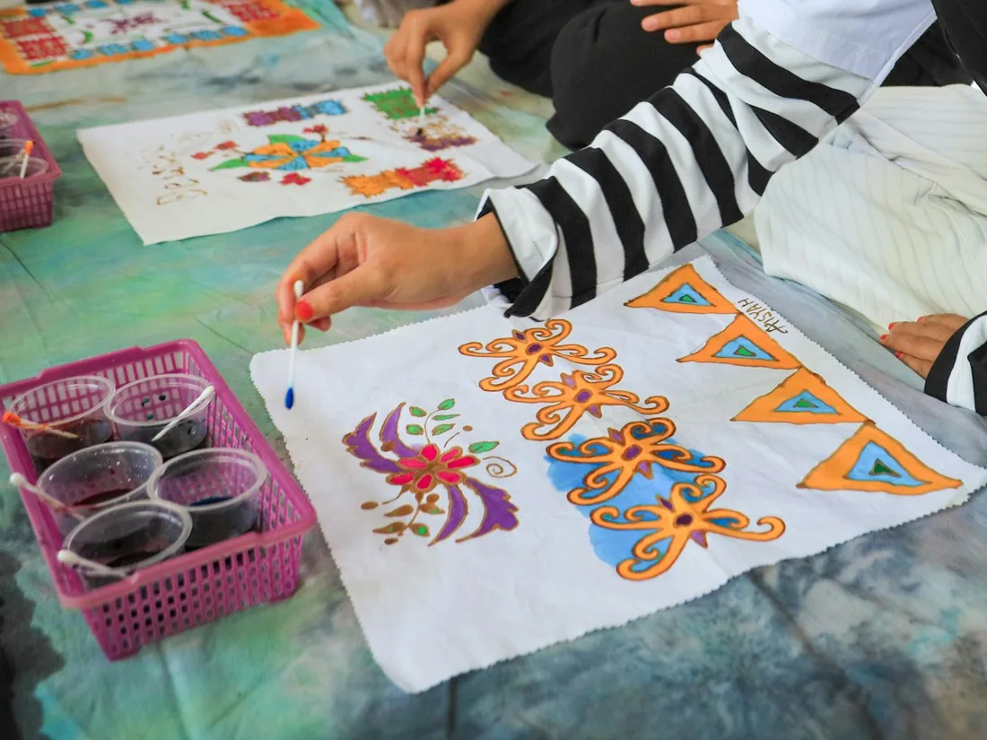 Manos aplicando color con pincel sobre un pañuelo de algodón dentro de las líneas de reserva, junto a vasos con tinte diluido